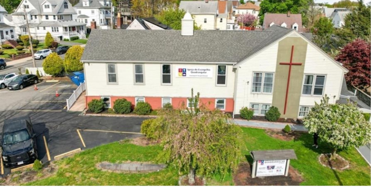 Aerial view of The Potters House Christian Fellowship in Massachusetts, showing a two-story beige church building with a red cross on the facade, surrounded by a residential neighborhood.